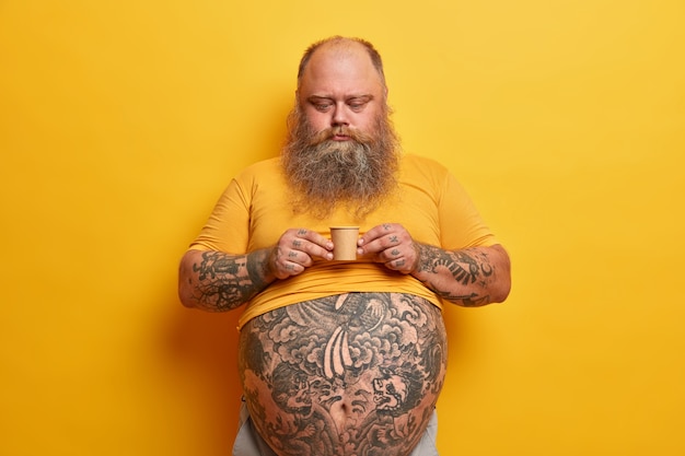 Homme barbu sérieux avec gros ventre, bras et ventre tatoués, tient une très petite tasse de café en carton contenant beaucoup de sucre, apprécie la boisson aromatique de caféine, porte un t-shirt jaune, pose seul à l'intérieur