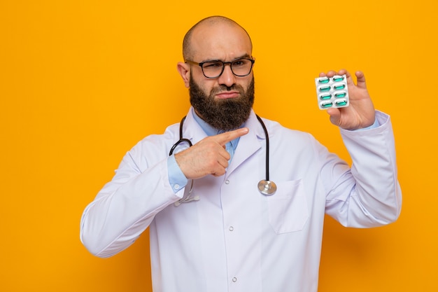 Homme barbu sérieux médecin en blouse blanche avec stéthoscope autour du cou portant des lunettes tenant un blister avec des pilules pointant avec l'index dessus