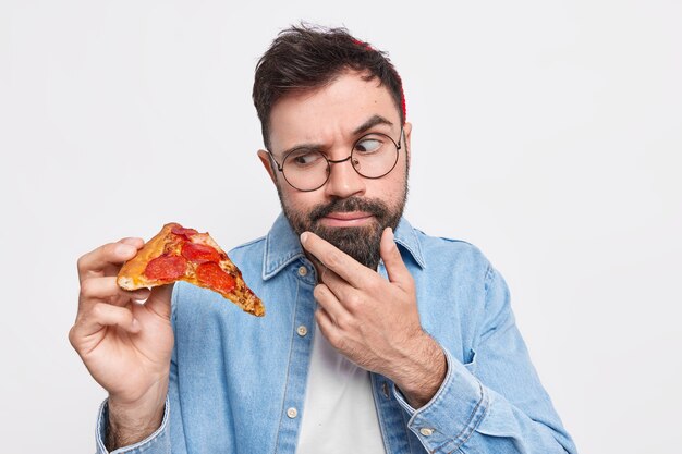 Un homme barbu sérieux regarde une tranche de pizza appétissante ressent la tentation de manger de la malbouffe tient le menton vêtu d'une chemise en jean porte des lunettes rondes