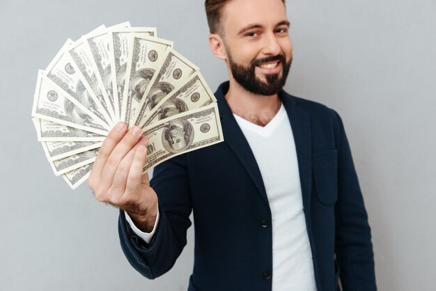 Homme barbu souriant dans des vêtements busines montrant de l'argent et regardant la caméra sur gris