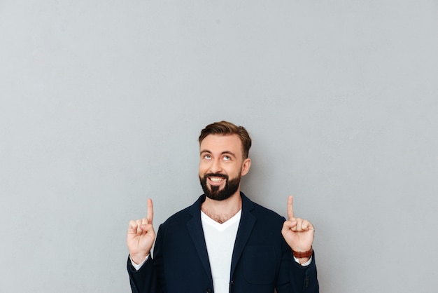 Homme barbu souriant dans des vêtements busines pointant et levant sur gris