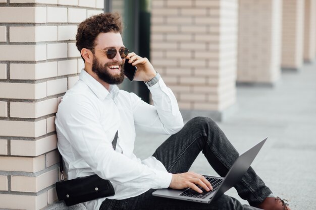 Un homme barbu, souriant et élégant assis sur la farine dans les rues de la ville près d'un centre de bureaux moderne et travaillant avec son ordinateur portable et son téléphone