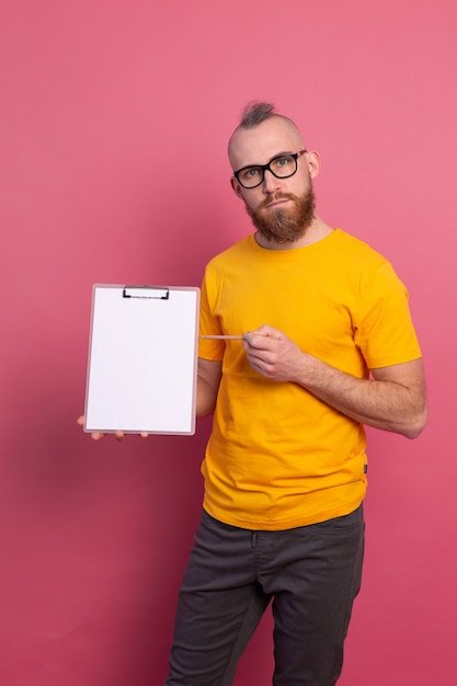 Homme barbu souriant portant des lunettes portant des vêtements décontractés tenant un presse-papiers avec un papier vierge pointant