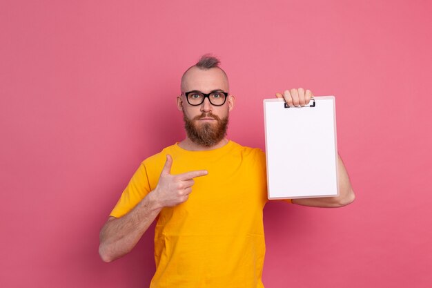 Homme barbu souriant portant des lunettes portant des vêtements décontractés tenant un presse-papiers avec un papier vierge pointant