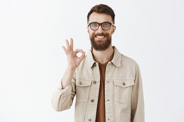 Homme barbu souriant satisfait dans des verres posant contre le mur blanc