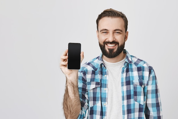 Homme barbu souriant satisfait montrant l'écran du smartphone, promotion de l'application