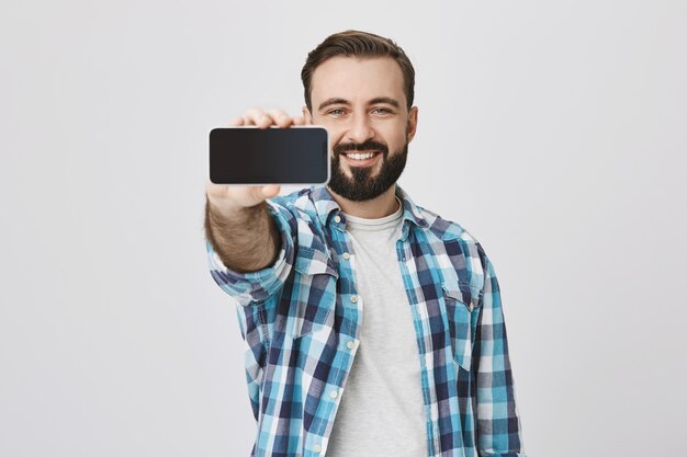 Homme barbu souriant satisfait montrant l'écran du smartphone, promotion de l'application