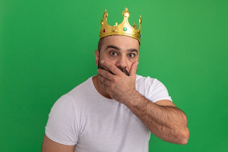 Photo gratuit homme barbu en t-shirt blanc portant une couronne d'or étant choqué couvrant la bouche avec la main debout sur le mur vert