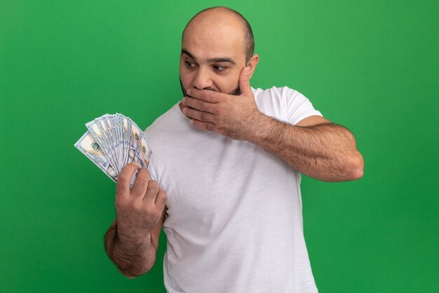 Photo gratuite homme barbu en t-shirt blanc tenant de l'argent à l'argent étonné et surpris couvrant la bouche avec la main debout sur le mur vert