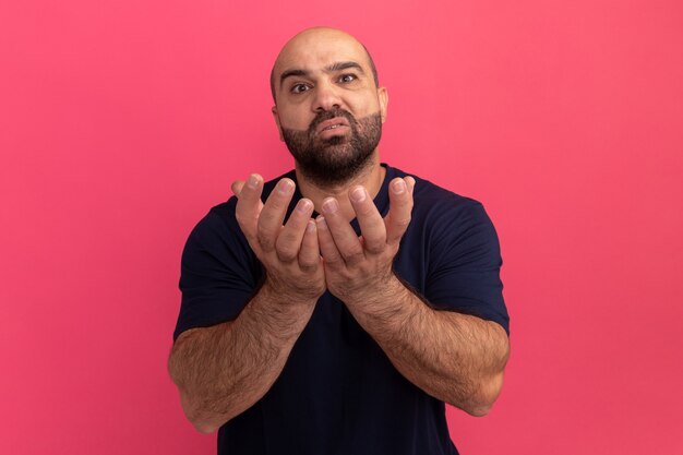 Homme barbu en t-shirt bleu marine avec les bras levés la mendicité avec une expression d'espoir debout sur un mur rose