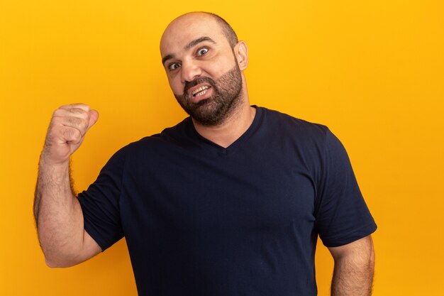 Homme barbu en t-shirt bleu marine confus pointant vers l'arrière debout sur un mur orange