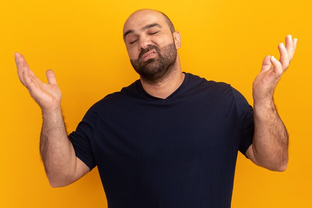 Homme barbu en t-shirt bleu marine à la confusion et incertain avec les bras levés debout sur un mur orange