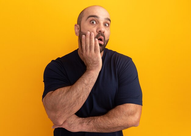 Homme barbu en t-shirt bleu marine étonné et surpris debout sur un mur orange