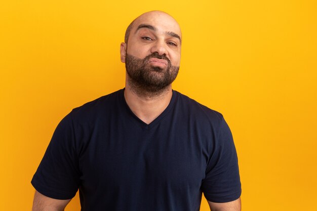 Homme barbu en t-shirt bleu marine avec une expression sérieuse et confiante debout sur un mur orange