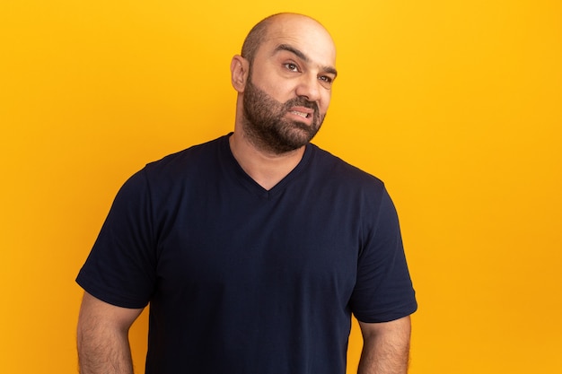 Homme barbu en t-shirt bleu marine faisant la bouche ironique avec une expression déçue debout sur un mur orange