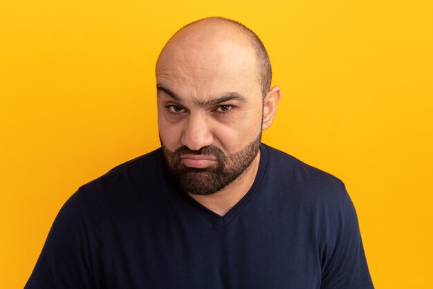 Homme barbu en t-shirt bleu marine faisant la bouche ironique avec une expression déçue debout sur un mur orange