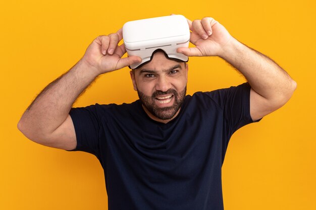 Homme barbu en t-shirt bleu marine avec des lunettes de réalité virtuelle avec une expression agacée debout sur un mur orange