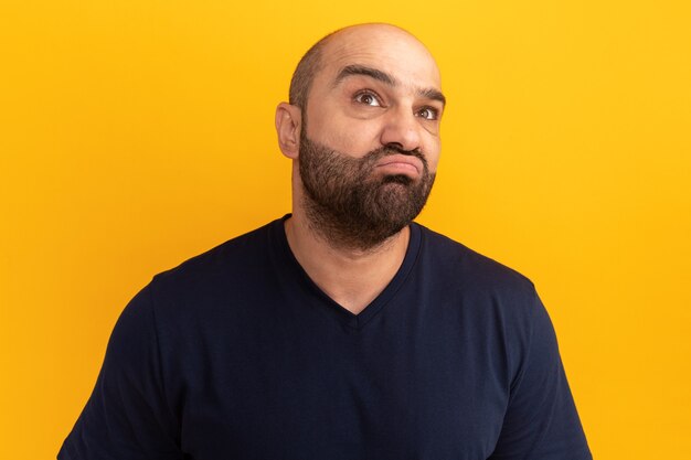 Homme barbu en t-shirt bleu marine à la perplexité debout sur un mur orange