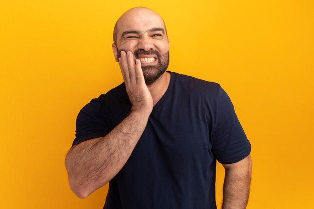 Homme barbu en t-shirt bleu marine à la recherche de mal à toucher sa joue ayant mal aux dents debout sur un mur orange