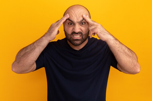 Homme barbu en t-shirt bleu marine avec visage en colère touchant sa tête debout sur un mur orange