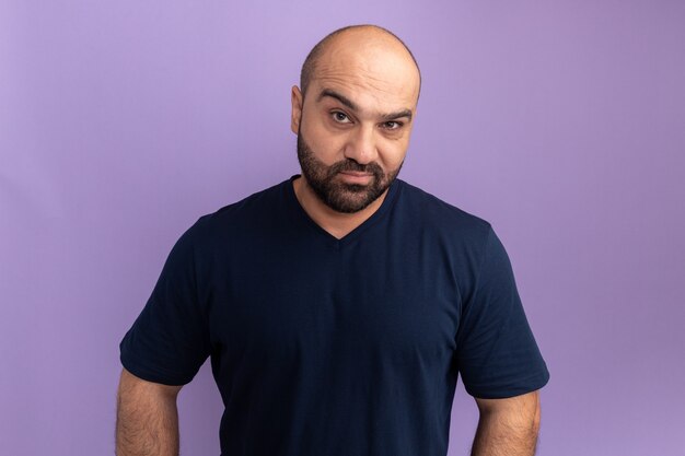 Homme barbu en t-shirt bleu marine avec visage sérieux debout sur un mur violet