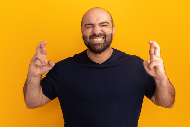 Homme barbu en t-shirt bleu marine avec les yeux fermés faisant voeu croisant les doigts debout sur le mur orange