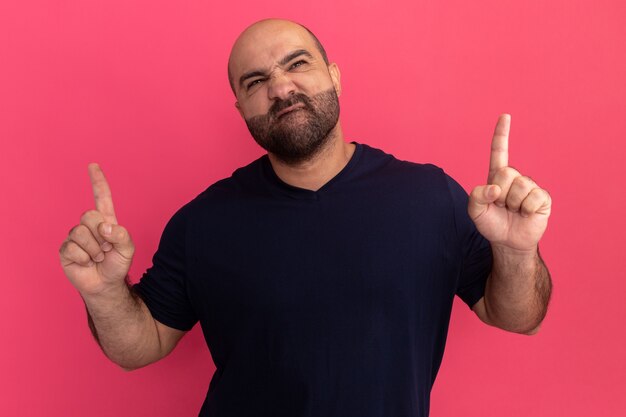 Homme barbu en t-shirt marine jusqu'à faire la bouche ironique avec une expression déçue montrant les index debout sur le mur rose