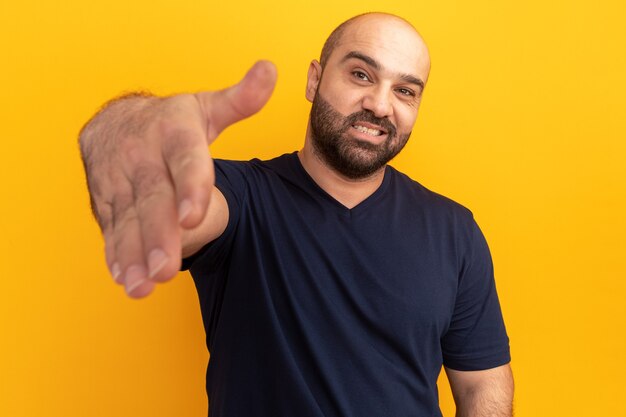 Homme barbu en t-shirt marine souriant offrant un geste de salutation à la main debout sur un mur orange