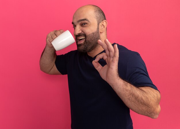 Homme barbu en t-shirt marine tenant une tasse de thé va boire heureux et positif montrant signe ok debout sur un mur rose