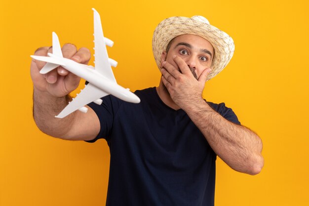 Homme barbu en t-shirt noir et chapeau d'été tenant un avion jouet étonné et inquiet couvrant la bouche avec la main debout sur le mur orange