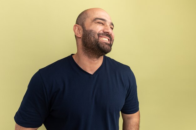 Homme barbu en t-shirt noir à côté souriant joyeusement heureux et positif debout sur mur vert