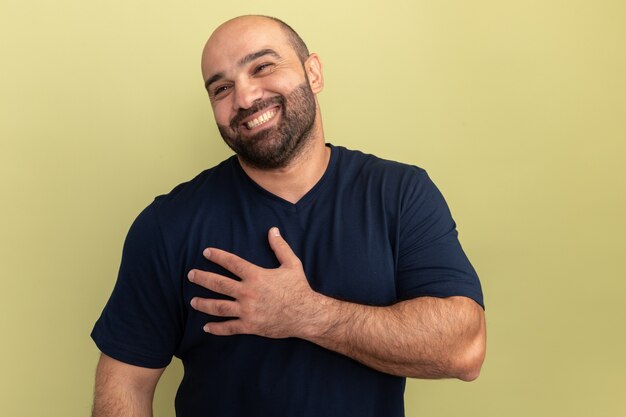 Homme barbu en t-shirt noir à côté souriant joyeusement tenant la main sur sa poitrine se sentant reconnaissant debout sur le mur vert