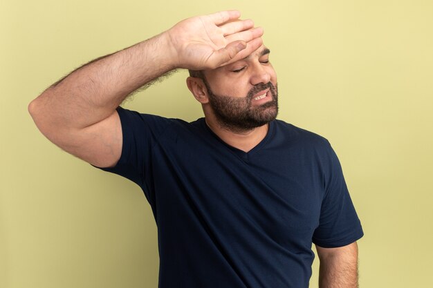 Homme barbu en t-shirt noir à la fatigue et agacé avec la main sur le front debout sur le mur vert