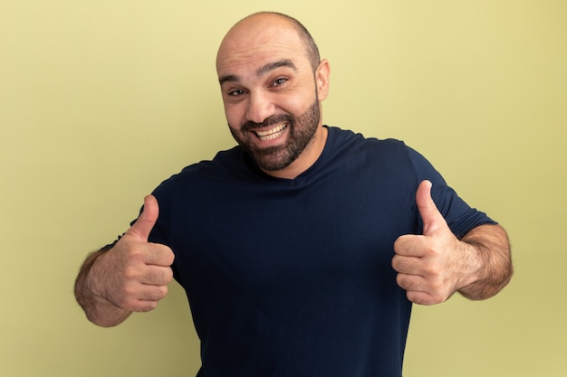 Homme barbu en t-shirt noir heureux et excité montrant les pouces vers le haut debout sur le mur vert