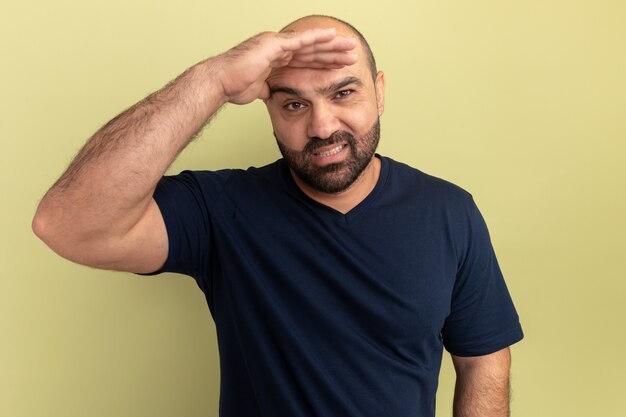 Homme barbu en t-shirt noir à la recherche de loin avec la main sur la tête souriant confus debout sur mur vert