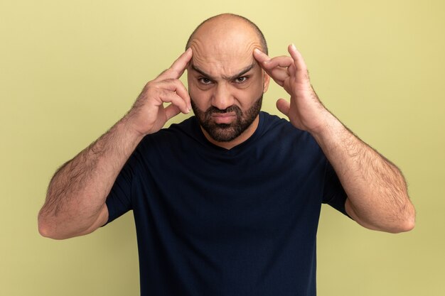 Homme barbu en t-shirt noir à la recherche de mal et ennuyé de toucher sa tête souffrant de forts maux de tête debout sur un mur vert