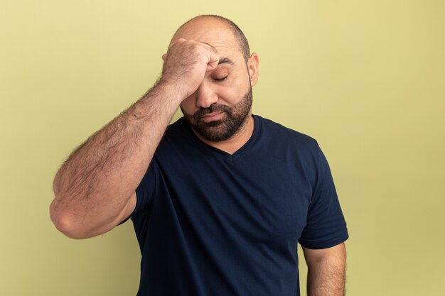 Homme barbu en t-shirt noir à la recherche de mal et ennuyé de toucher sa tête souffrant de forts maux de tête debout sur un mur vert