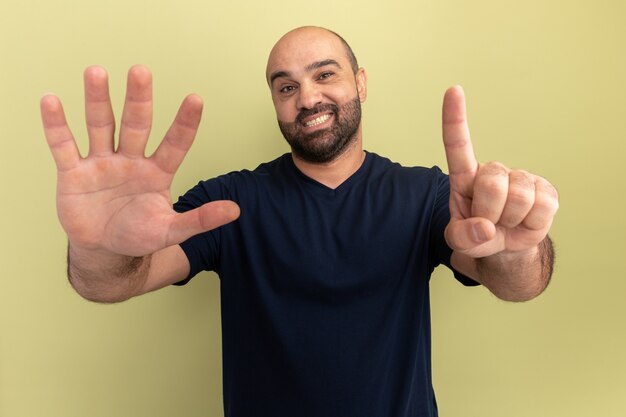 Homme barbu en t-shirt noir souriant montrant le numéro six debout sur un mur vert