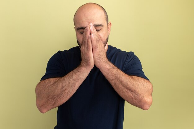 Homme barbu en t-shirt noir tenant les mains ensemble sur son visage déprimé et inquiet debout sur le mur vert