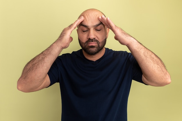 Homme barbu en t-shirt noir touchant sa tête à la souffrance souffrant de maux de tête debout sur mur vert