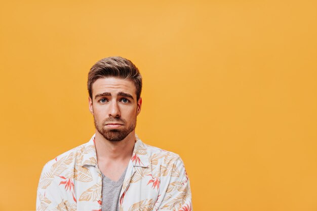 Homme barbu triste aux yeux bleus en chemise imprimée à la mode et t-shirt gris regardant dans la caméra sur un mur orange isolé