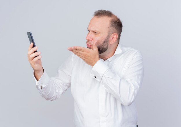 Homme barbu vêtu d'une chemise blanche tenant un smartphone regardant l'écran ayant un appel vidéo soufflant un baiser debout sur un mur blanc