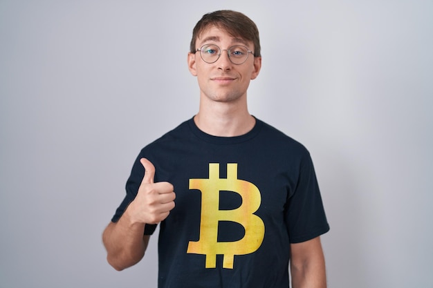 Photo gratuite homme blond caucasien portant un t-shirt bitcoin faisant un geste de pouce levé heureux avec une expression d'approbation de la main regardant la caméra montrant le succès