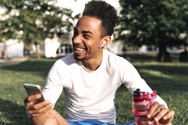 Homme de bonne humeur à la peau foncée bouclée en t-shirt blanc à manches longues et short bleu rit sincèrement, tient un téléphone et une bouteille d'eau rose dans le parc