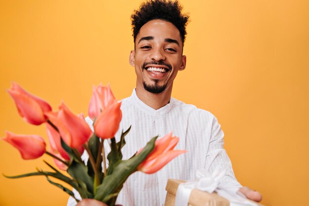 Homme de bonne humeur présente des fleurs et des cadeaux Portrait d'un mec brune en chemise blanche tenant un bouquet de tulipes roses sur fond orange