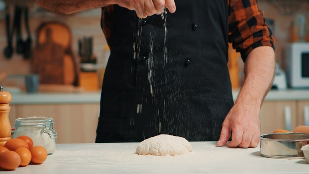 Homme de boulangerie senior tamisant la farine de blé sur la pâte à pétrir. Chef âgé à la retraite avec bonete et saupoudrage uniforme, tamisage étalant des ingrédients rew avec cuisson à la main de pizza et de pain faits maison.