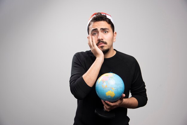 Homme bouleversé au chapeau du père Noël posant avec un globe.