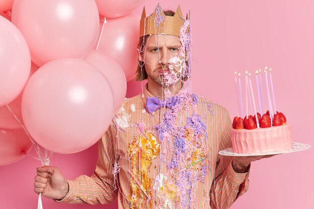 Joyeux Anniversaire Homme Avec Une Expression Heureuse Porte Des Vetements De Fete Sales Couronne De Papier Tient Le Gateau Et Les Ballons Pose A La Fete Contre Le Mur Rose Celebre L Anniversaire
