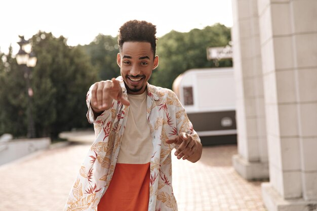 Homme brun bouclé à la peau foncée en t-shirt de couleur et chemise à fleurs élégante souriant et pointant vers la caméra à l'extérieur