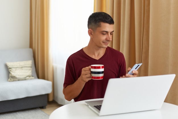 Homme brun séduisant portant un t-shirt de style décontracté bordeaux assis à table avec un ordinateur portable, utilisant un téléphone intelligent pour consulter ses e-mails ou les réseaux sociaux, savourant une boisson chaude le matin.